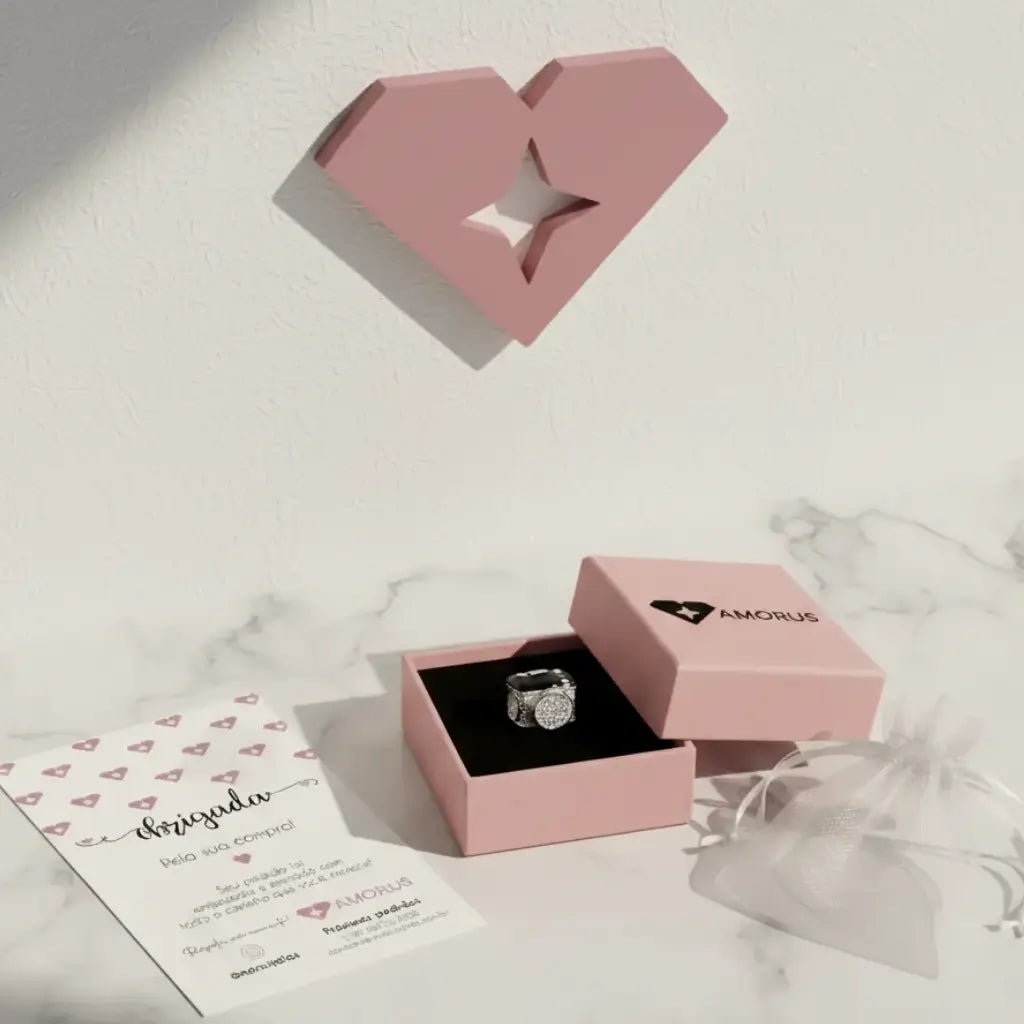 Berloque em formato de câmera fotográfica apresentado dentro de uma caixa rosa da marca AMORUS, acompanhado de cartão de agradecimento e saquinho de tecido branco.