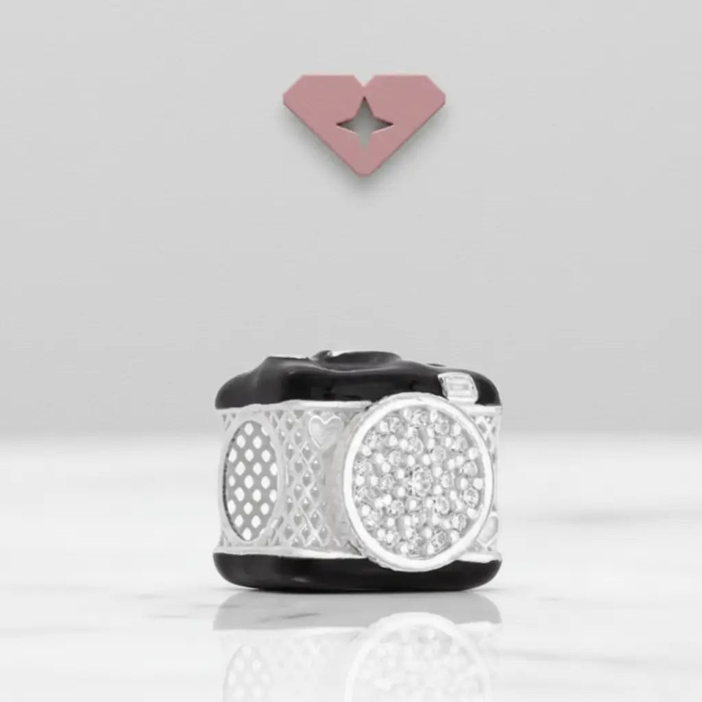 Berloque em formato de câmera fotográfica exposto em fundo branco com logo em formato de coração ao fundo, destacando os detalhes cravejados na lente.
