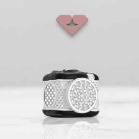 Berloque em formato de câmera fotográfica exposto em fundo branco com logo em formato de coração ao fundo, destacando os detalhes cravejados na lente.