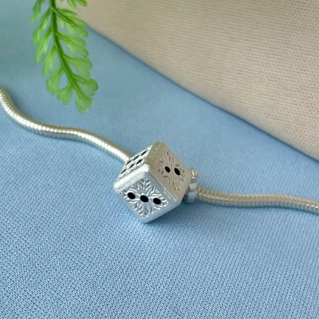 Berloque dado em Prata 925 exibido sobre fundo azul, com detalhe de planta decorativa
