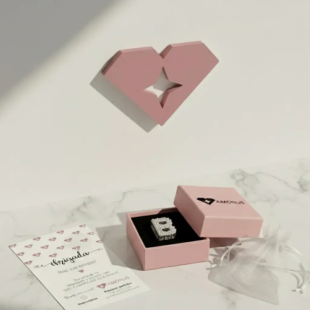 Berloque letra B cravejado com zircônias, exibido em uma caixa rosa da marca AMORUS, com cartão de agradecimento e saquinho de tule ao lado