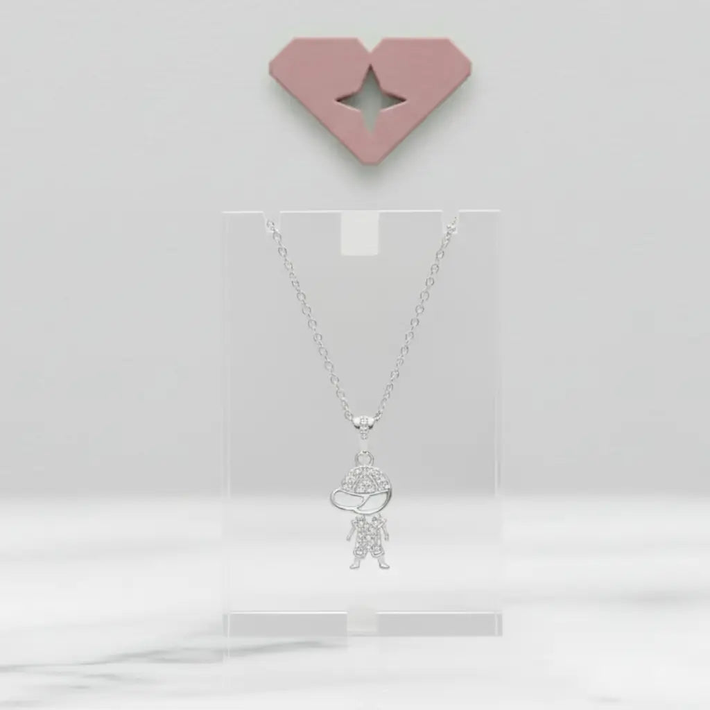 Colar de prata com pingente de menino cravejado em exibição sobre suporte acrílico transparente, com fundo minimalista e detalhe decorativo de diamante rosa na parede.