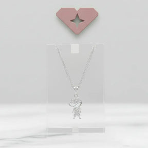 Colar de prata com pingente de menino cravejado em exibição sobre suporte acrílico transparente, com fundo minimalista e detalhe decorativo de diamante rosa na parede.