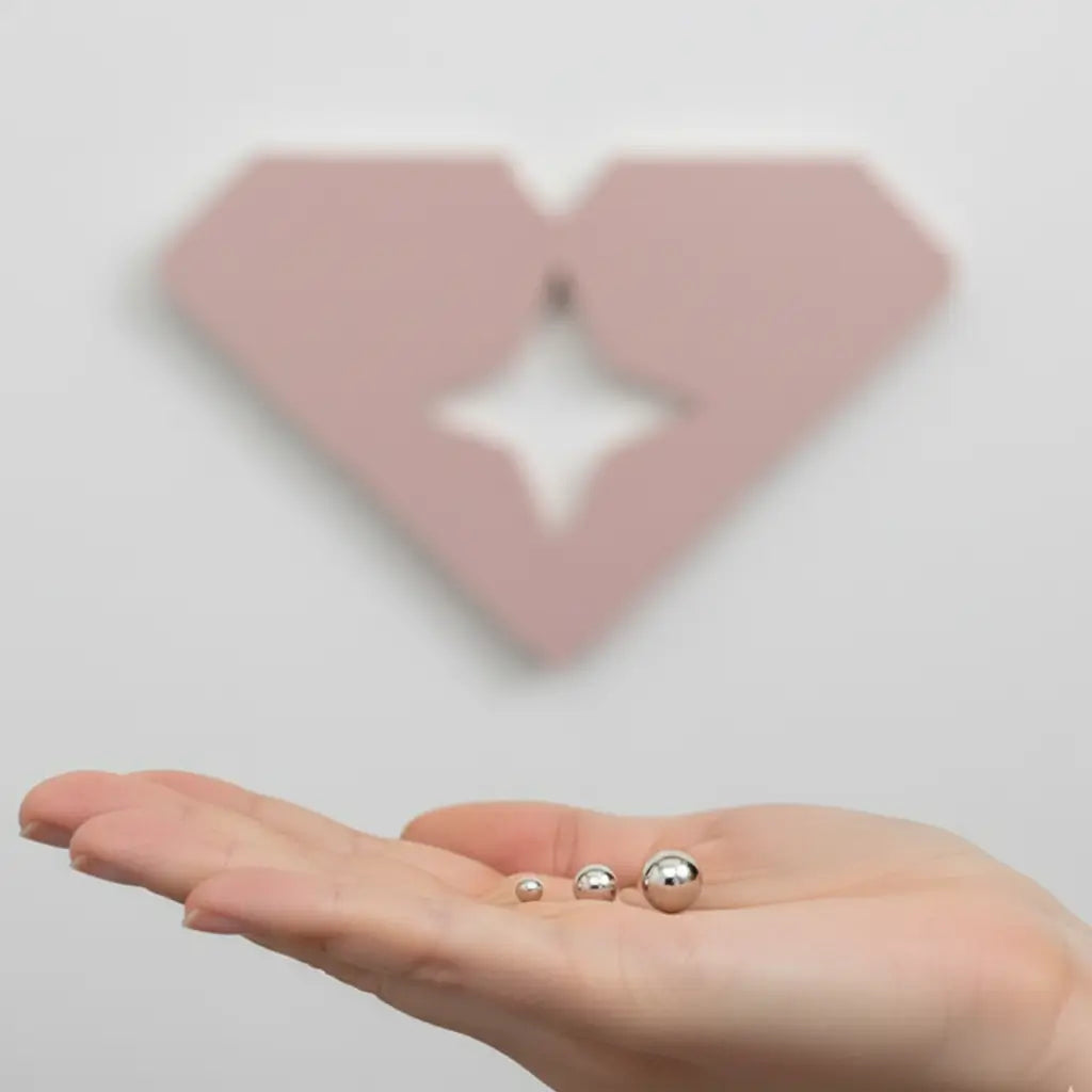 Trio de brincos bolinha em aço inox na palma da mão, com logo rosa da marca em segundo plano.